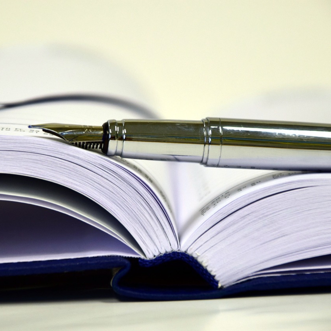 Open book with a blue cover and a silver fountain pen resting on its pages. The image conveys a sense of writing, creativity, and contemplation.