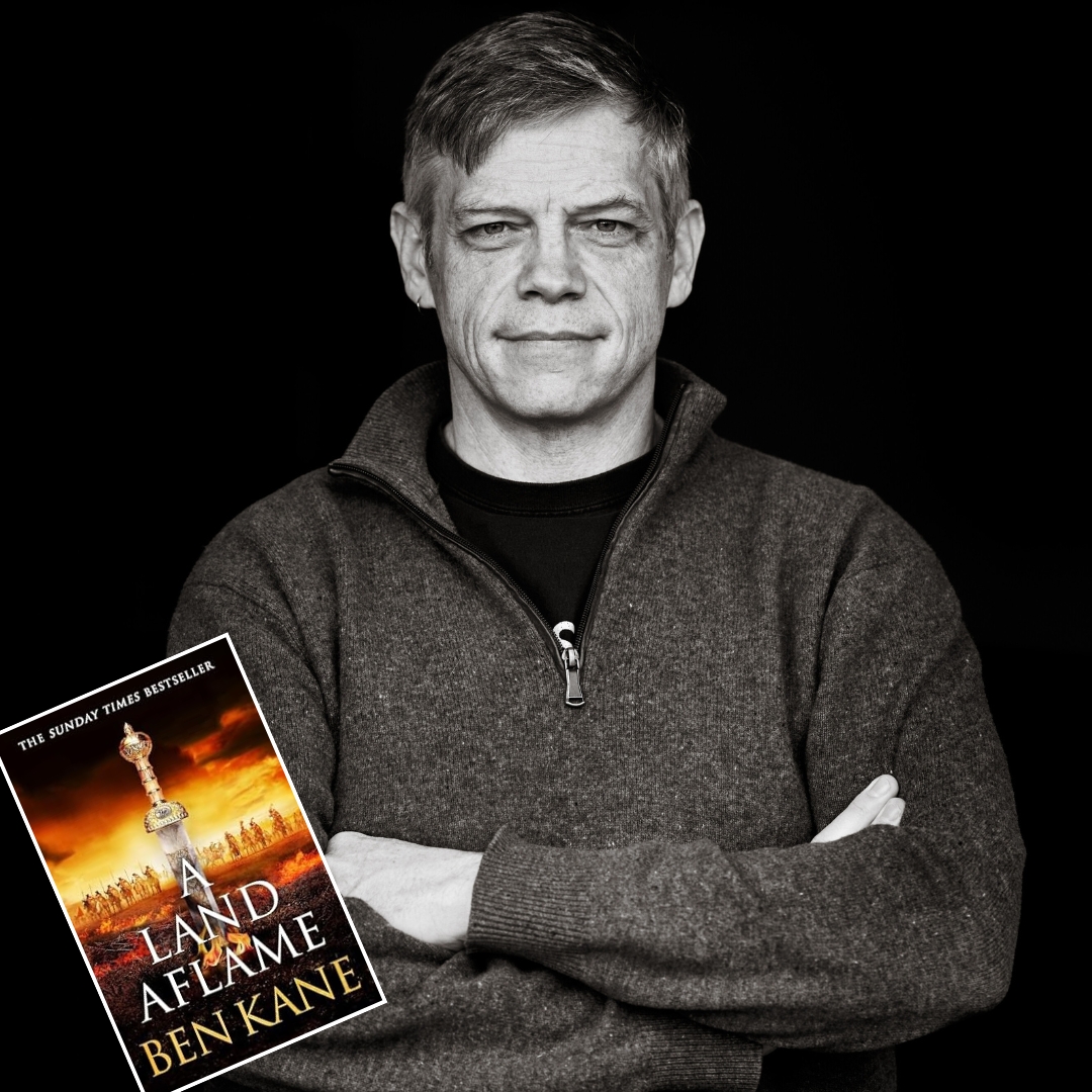 A grayscale image of a person with crossed arms, wearing a sweater. A book titled "A Land Aflame" by Ben Kane is positioned in the foreground.