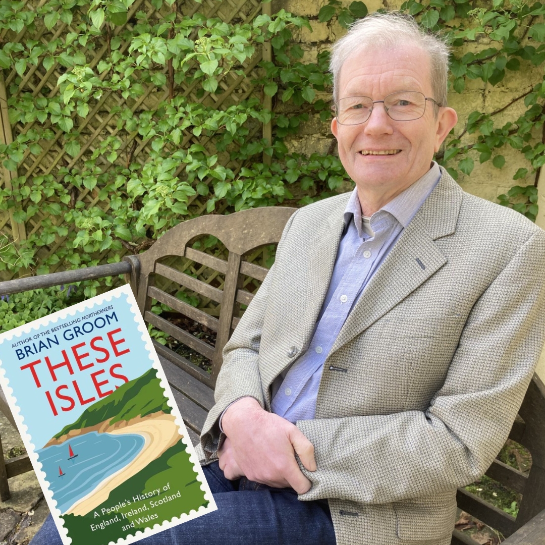 A man in a checked blazer sits on a wooden bench against a leafy green background, smiling. A book titled "These Isles" with a coastal illustration is in view.