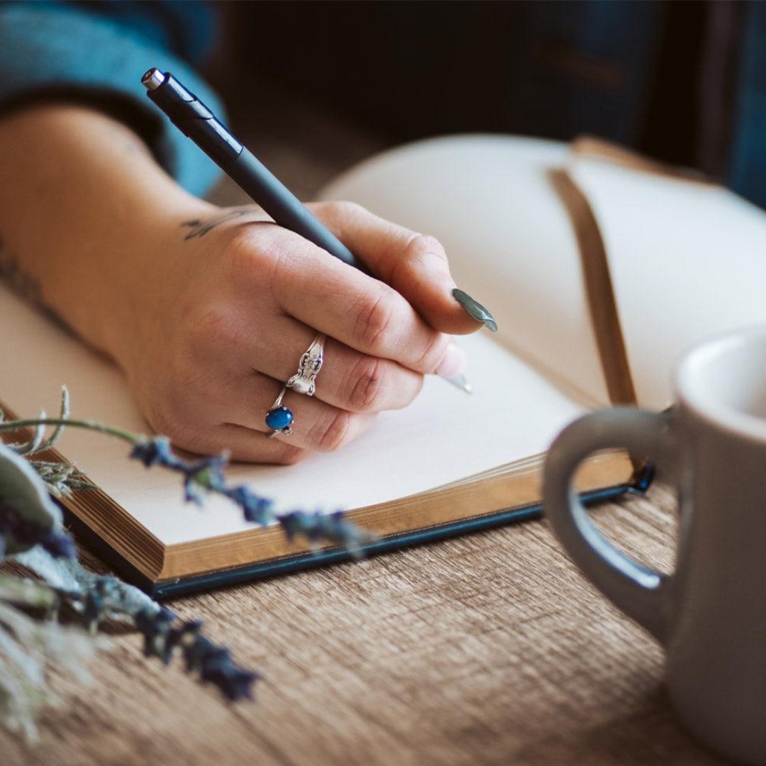 A hand with rings holds a pen, writing in an open notebook. A coffee mug sits nearby on a wooden table, with lavender flowers in the foreground. Calm and focused atmosphere.