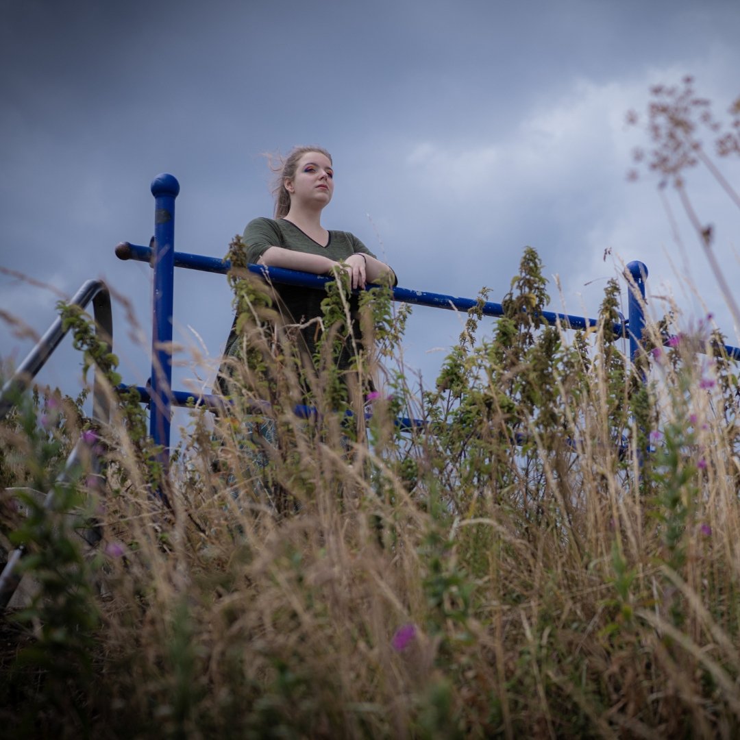A woman in a green shirt stands pensively behind a blue railing, surrounded by tall grass and wildflowers, against a cloudy, moody sky.